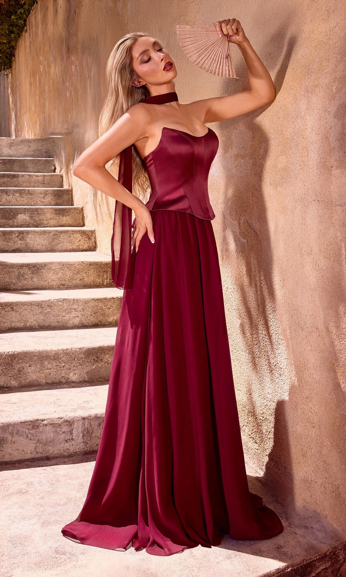 A woman in the Ladivine CD386 Strapless Long Corset Prom Dress stands on stone steps, holding a fan near her face with one hand and resting the other on her hip against a textured wall.
